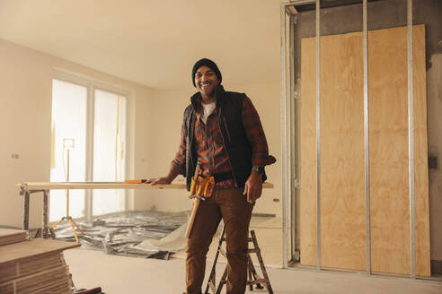 Ein glücklicher Bauunternehmer mit einem Werkzeuggürtel steht in der Küche eines Hauses, das gerade renoviert wird, und blickt lächelnd in die Kamera. Der Bauunternehmer arbeitet an der Umgestaltung des Innenraums und ist mit Bauwerkzeugen ausgestattet, um das Haus zu modernisieren. - JLPSF30958