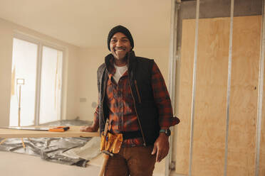 Happy DIY enthusiast wearing a tool belt and standing in a kitchen during renovations. He confidently looks at the camera, ready to upgrade and remodel his home with his carpenty expertise. - JLPSF30960