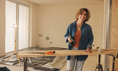 A joyful female contractor works on remodeling an interior kitchen. She holds a drill tool, happily refurbishing the wood baseboards. Her expertise and dedication shine through as she adds a personal touch to the home improvement project. - JLPSF30964