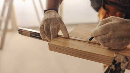 Ein männlicher Zimmermann mit Arbeitshandschuhen misst und markiert ein Holzbrett in einem Haus. Dieser Facharbeiter bringt sein Fachwissen ein, um das Innere des Hauses zu verschönern. Er konzentriert sich auf Verkleidungsarbeiten und zeigt sein handwerkliches Können. - JLPSF30971