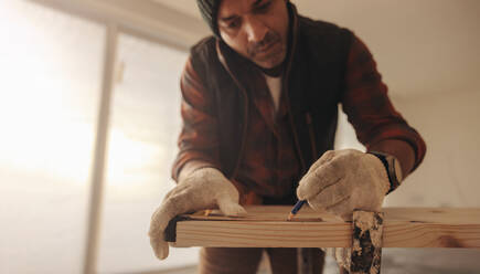 Geschickter Bauunternehmer bei der Innenrenovierung eines Hauses. Er arbeitet in einem Innenraum, insbesondere in der Küche. Er markiert mit einem Bleistift die Maße auf dem Holz, um präzise Schreiner- und Verkleidungsarbeiten durchzuführen. - JLPSF30973