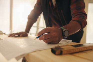 Ein Bauarbeiter markiert Baupläne mit einem Bleistift und beweist damit sein Geschick und seine Professionalität. Die Innenumgebung deutet auf ein Heimwerkerprojekt hin, das möglicherweise einen Bau oder eine Renovierung beinhaltet. - JLPSF30978