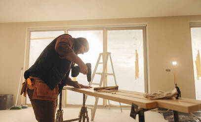 Generalunternehmer bei der Renovierung einer Küche in einem Haus. Ein Bauarbeiter verwendet eine Bohrmaschine, um Holzbretter für die Installation von Sockelleisten zu bearbeiten. Ein geschickter Arbeiter und Werkzeuge sorgen für eine erfolgreiche Innenrenovierung. - JLPSF30985