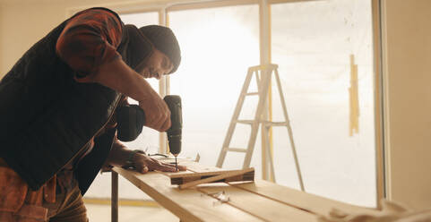 Bauarbeiter, der ein Haus mit Handwerkzeugen renoviert. Mit einer Bohrmaschine befestigt er ein Holzbrett für die Verkleidung in der Küche. Bauarbeiter, der ein Haus mit individuellen Holzarbeiten umgestaltet. - JLPSF30986