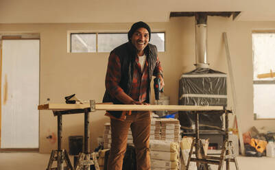 Älterer männlicher Arbeiter mit einem fröhlichen Lächeln, in einer Küche. Er renoviert und verschönert die Inneneinrichtung, wobei er sich auf Fußleisten und Holzarbeiten konzentriert. Mit einer Bohrmaschine zeigt er sein Fachwissen im Bereich Bau und Heimwerken. - JLPSF30990