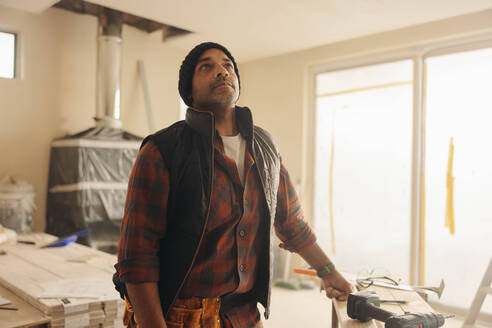 Ein nachdenklicher Bauarbeiter mit Werkzeuggürtel steht in einem Wohnzimmer und denkt über Renovierungsideen nach. Dieser reife Bauunternehmer widmet sich der Aufwertung und Renovierung des Hauses mit seinen Fähigkeiten. - JLPSF30993