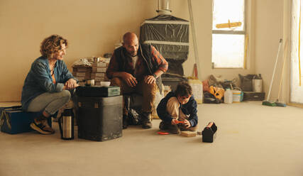 Parents on a home renovation project take a coffee break while their son plays with the tools. They work together as a team, embracing the challenges of DIY home improvement with smiles. - JLPSF31013