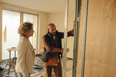 Construction worker and a homeowner collaborate on a home renovation project. They discuss plans indoors near a doorframe, working together to remodel and redesign the interior of the house. - JLPSF31025