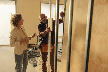 Ein männlicher Umgestalter und eine Architektin besprechen die Pläne für ein Hausrenovierungsprojekt. Sie stehen in der Nähe eines Türrahmens im Inneren des Hauses und planen die Umgestaltung der Türen mit einer Holzprobe. - JLPSF31031