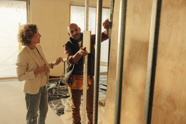 Male remodeler and a female architect are discussing plans for a home renovation project. They stand near a doorframe in the house's interior, planning the remodel of the doors with a wood sample. - JLPSF31031