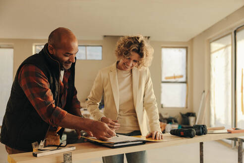 Ein männlicher Bauunternehmer und eine Hausbesitzerin unterzeichnen in einem Haus einen Vertrag. Das Dokument symbolisiert das Fachwissen des Bauunternehmers im Bereich der Hausrenovierung. Sie lächeln und zeigen damit ihre Zufriedenheit mit der Zusammenarbeit. - JLPSF31037
