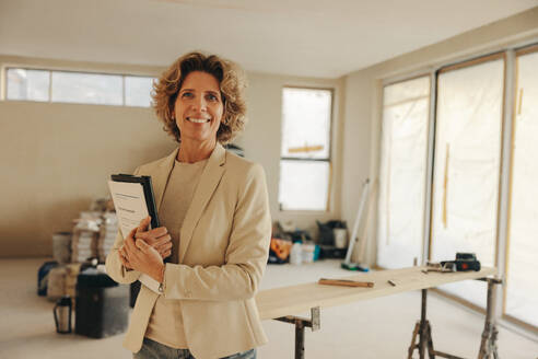 Happy female homeowner stands in her kitchen, holding paperwork for a home renovation. With a smile, she confidently looks at the camera, showcasing her involvement in the ongoing construction. - JLPSF31044