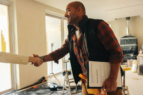 Ein glücklicher, reifer Bauunternehmer schüttelt einem Hausbesitzer die Hand, nachdem er den Papierkram unterschrieben hat, mit dem ein Renovierungsprojekt abgeschlossen wurde. - JLPSF31047
