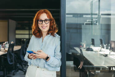 Portrait of a confident, smiling businesswoman in glasses standing in her office, holding a smartphone. This professional redhead embodies the modern workplace and the lifestyle of a successful worker. - JLPSF31052