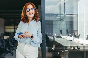 Smiling businesswoman with ginger hair stands in her office, wearing glasses and holding a smartphone. She is a professional worker and entrepreneur, happily using technology for her business. - JLPSF31053