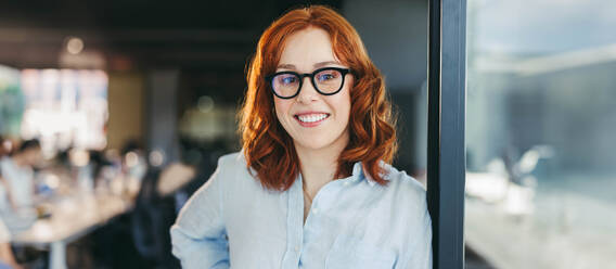 Confident redhead business woman with glasses stands in a panoramic office, happy and successful. She is a dedicated employee in a tech company, embodying the ideal lifestyle of a professional woman. - JLPSF31055