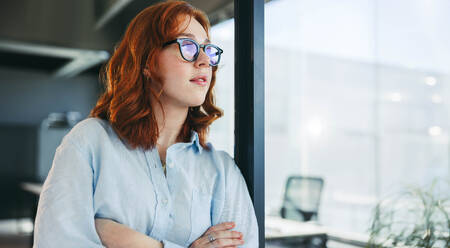Ginger-haired businesswoman stands confidently in an office, arms crossed, contemplating her work. With glasses on, she embodies professionalism and is a valuable asset to any company. - JLPSF31057