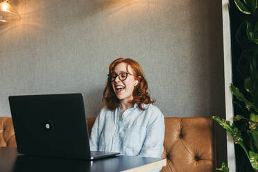 Professional woman with ginger hair works happily in a cozy cafe-like office. Engaged in a video call, she smiles while using her laptop. This image showcases remote work and the modern workplace. - JLPSF31059