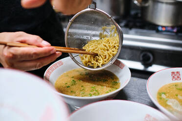 Crop anonymous chef with chopsticks putting noodles in bowl with bouillon while serving ramen soup on table in kitchen - ADSF48867
