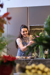 Professional female florist in uniform smiling and looking down while arranging bouquet of blooming flowers during workday in modern floral shop - ADSF48868