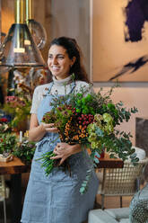 Smiling young woman in blue apron touching arranged flower bouquet and looking away while working in floral shop on blurred background - ADSF48869