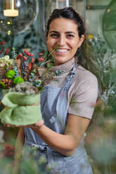 Cheerful woman in blue apron smiling and looking at camera while working in floral shop and demonstrating arranged bouquet of blooming flowers - ADSF48870