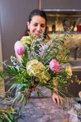 Smiling woman in apron standing at marble table with blooming flowers arranged in bouquet in modern floral shop - ADSF48871