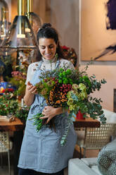Professional female florist in blue apron smiling and looking down at arranged bouquet of blossoming flowers while working in floral shop - ADSF48873