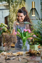 Professional female florist in apron standing at table with various plants and flowers and looking at blossoming carnation while working in floral shop - ADSF48875