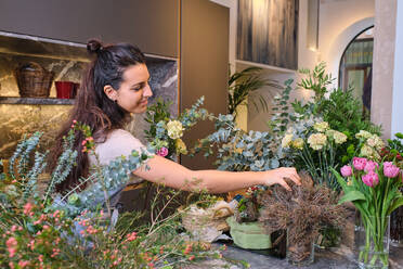 Smiling female florist in apron looking at blossoming flowers and arranging bouquet in modern floral shop - ADSF48877