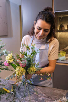 Smiling woman in blue apron standing at table and looking at bright blossoming flowers while arranging bouquet in floral shop - ADSF48878