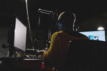 Back view of anonymous female radio host in headphones mixing audio while performing live broadcast in dark studio - ADSF48888