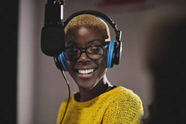Optimistic ethnic female radio host in bright yellow sweater headphones and glasses recording podcast near microphone in studio - ADSF48902