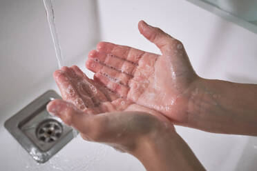 Hands of crop faceless kid washing hands with foamy soap in sink with water stream during hygienic routine in bathroom - ADSF48921