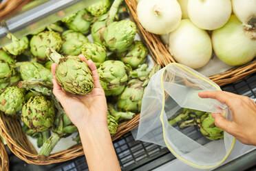 High angle of crop anonymous client choosing ripe artichoke and putting in eco friendly reusable bag while shopping in grocery store - ADSF48938