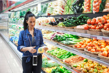 Ethnic lady standing near shelves with assorted vegetables in grocery store and buying fresh pepper in plastic bag - ADSF48940