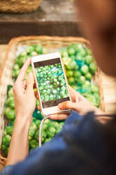 From above of crop unrecognizable person with mobile phone taking picture of artichoke pile in wicker basket in market - ADSF48941