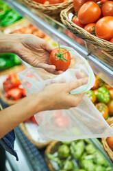 Crop faceless buyer putting ripe red tomato into ECO friendly nylon bag while standing near stall with various vegetables in supermarket - ADSF48942