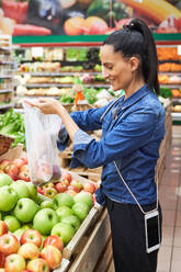 Side view of satisfied ethnic lady selecting fresh apples into plastic bag on blurred background in supermarket - ADSF48946