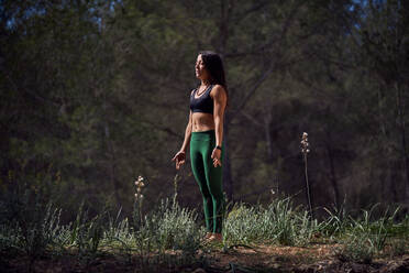 Full body of calm young Hispanic lady with long dark hair in leggings and crop top standing in forest with closed eyes while meditating in Mountain asana during yoga session - ADSF48950