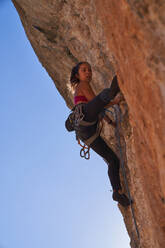 From below of female alpinist wearing safety harness sportswear and boots climbing steep cliff using dynamic rope carabines and chalkbag and looking away - ADSF48953