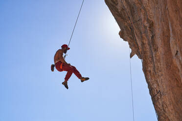 Full body side view of strong shirtless male alpinist hanging on rope while descending from rocky cliff against blue sky - ADSF48955