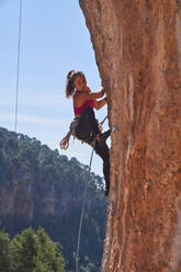 From below female alpinist wearing sportswear climbing boots and harness attached to dynamic rope climbing rocky mountain using quickdraws and looking down - ADSF48956