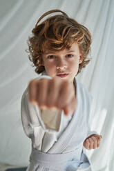 Confident preteen kid with curly hair in white kimono standing near curtain with clenched fists and punching during judo training - ADSF48960