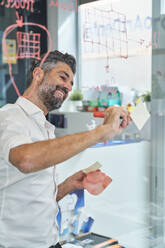 Through window of delighted male engineer taking notes on glass wall while working on design on face mask in light office - ADSF48973