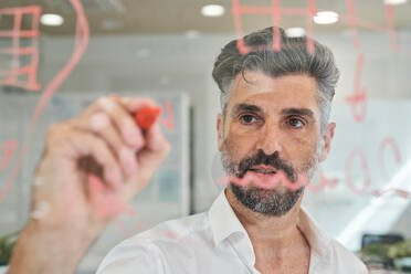 Through glass view of unshaven Hispanic man in shirt writing information on glass wall and working at office - ADSF48977