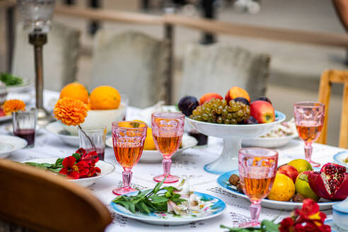 A vibrant outdoor table setting featuring an assortment of fresh fruits, colorful glasses filled with drinks, and decorative flowers placed on a white tablecloth. - ADSF48989