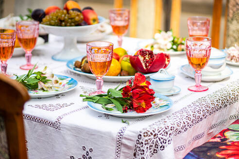 Brightly colored dining table set-up with pink beverages in crystal glasses, a plate showcasing red flowers, assorted fruits, and ornate porcelain dishes, all placed on a lace-edged white tablecloth. - ADSF48990