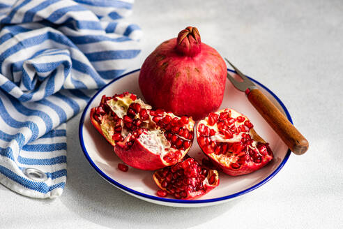 A fresh ripe pomegranate sits on a white and blue plate, surrounded by its red seeds and a rustic knife with a wooden handle. A striped napkin blue and white against gray background. - ADSF48996