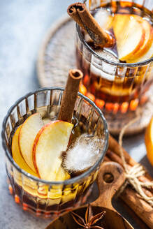 Fresh apple cider cocktails garnished with cinnamon sticks, star anise, and apple slices in a crystal glass filled with ice cubes on a textured background - ADSF48997